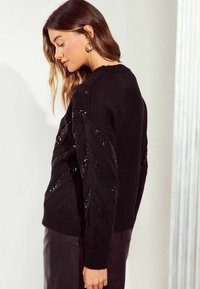 Maglione nero lavorato a maglia con un motivo testurizzato e paillettes, polsini a coste e design a girocollo. Indossato con una gonna plissettata.