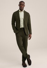 Traje verde oliva con chaqueta a medida y pantalones ajustados, combinado con una camisa verde clara. Unos zapatos de ante marrón completan el look.