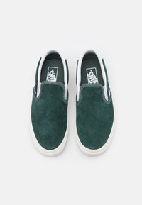Ciemnozielone zamszowe buty wsuwane z białymi gumowymi podeszwami i detalami. Wnętrze wykończone jest materiałem typu canvas, a na języku umieszczono logo marki.