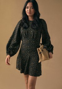 Sort kjole med gyldne blomsterprint, puffede ærmer og en figursyet talje, stylet med en beige ruskinds taske med mørke stropper.