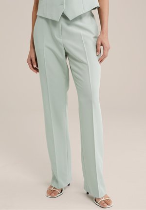 Femme portant un pantalon large sur mesure vert clair associé à des sandales blanches à talons ouvertes, debout devant un fond neutre.