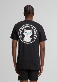 Svart bomull T-skjorte med et rundt grafisk trykk av en tegneserie-hvit katt og teksten "SLUTT Å VÆRE EN PUSSY" i fete hvite bokstaver.