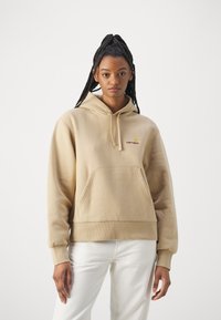 Beige hoodie with a kangaroo pocket, drawstring hood, ribbed cuffs, and a small embroidered logo on the chest. Made of soft, textured fabric.