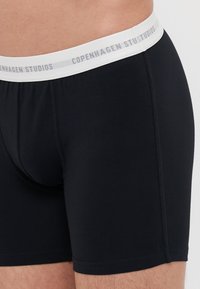 Čierne boxerky s bielym elastickým pásom, na ktorom je text "Copenhagen Studios" v šedej farbe. Vyrobené z mäkkého, pružného materiálu.