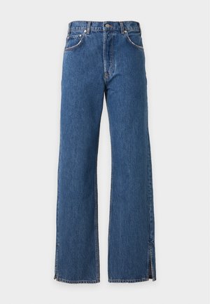 Frau mit langen dunklen Haaren, die ein graues T-Shirt mit Grafik, blaue Jeans mit Knöchelschlitzen und schwarze spitze High Heels trägt, stehend vor weißem Hintergrund.