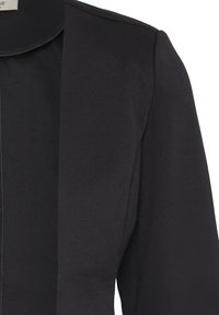 Blazer noir avec épaule structurée et col châle sur fond blanc.