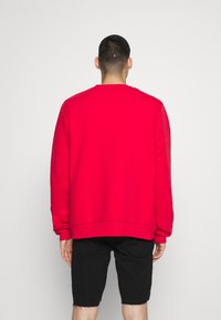 Felpa rossa con scollo a girocollo, polsi e orlo a costine. Il tessuto appare liscio, indossata su pantaloni di jeans neri a pantaloncino, vista da dietro.