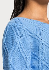 Lähikuva henkilöstä, joka on pukeutunut vaaleansiniseen neuleeseen, jossa on teksturoitu geometrinen kuvio ja letitetty hius.