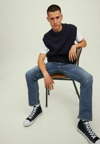 Maglione senza maniche a coste blu navy sopra una t-shirt bianca, jeans blu e sneakers alte nere, seduto su una sedia di legno contro uno sfondo chiaro.