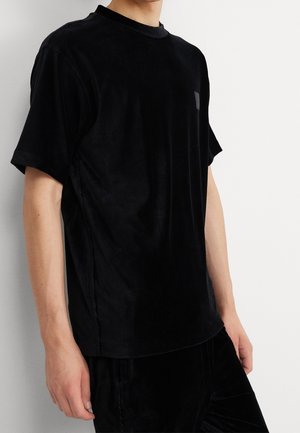 Person iført en kortærmet sort fløjls-T-shirt mod en ensfarvet lys baggrund, der viser overkrop og arm.