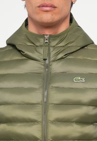 Jachetă puffer, matlasată, de culoare verde măsliniu, cu fermoar frontal și glugă atașată. Prezintă un mic logo Lacoste pe partea stângă a pieptului.