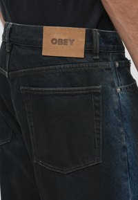 Temno modri kavbojki imajo rjavo usnjeno označbo z blagovno znamko "OBEY" na hrbtni strani. Tkanina pokaže kontrastne odtenke in vidne podrobnosti šivanja.