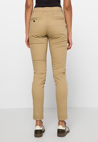 Tan slim-fit pants with two back pockets and a single button detail. Made of soft fabric with a smooth texture. Worn with dark sneakers.