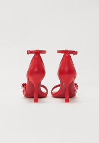 Chaussures à talons hauts rouges avec une finition en cuir lisse, une bride ajustable à la cheville et un design ouvert à l'avant, avec un nœud décoratif sur le côté.