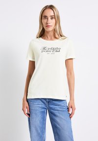 T-shirt en coton beige clair avec un col rond, des manches courtes et un texte noir "The infinite palm Club EST. 2025" sur le devant.