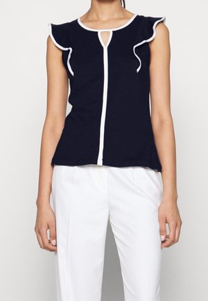 Femme portant un haut bleu marine sans manches avec un volant blanc et un décolleté en forme de trou de serrure, assorti à un pantalon blanc taille haute.