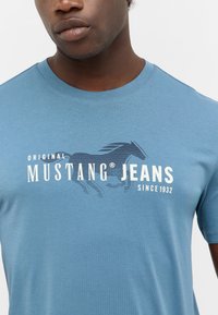 Niebieska koszulka z krótkim rękawem, wykonana z miękkiego materiału, z białym wzorem graficznym przedstawiającym konia oraz napisem "MUSTANG JEANS".