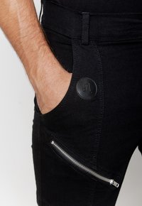 Czarne szorty dżinsowe z boczną kieszenią na zamek, skórzaną łatą z logo i kontrastowym szwem. Materiał ma gładką teksturę.