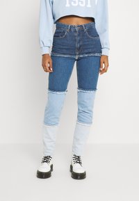 Jeans de mezclilla con un diseño de patchwork que combina tonos de azul oscuro y claro. Presentan bordes deshilachados y un corte de cintura alta. Combinados con botas blancas.