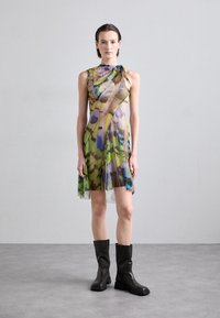 Robe multicolore avec une superposition transparente, présentant un motif floral dans des teintes de vert, bleu et brun. Associée à des bottes noires jusqu'aux genoux.