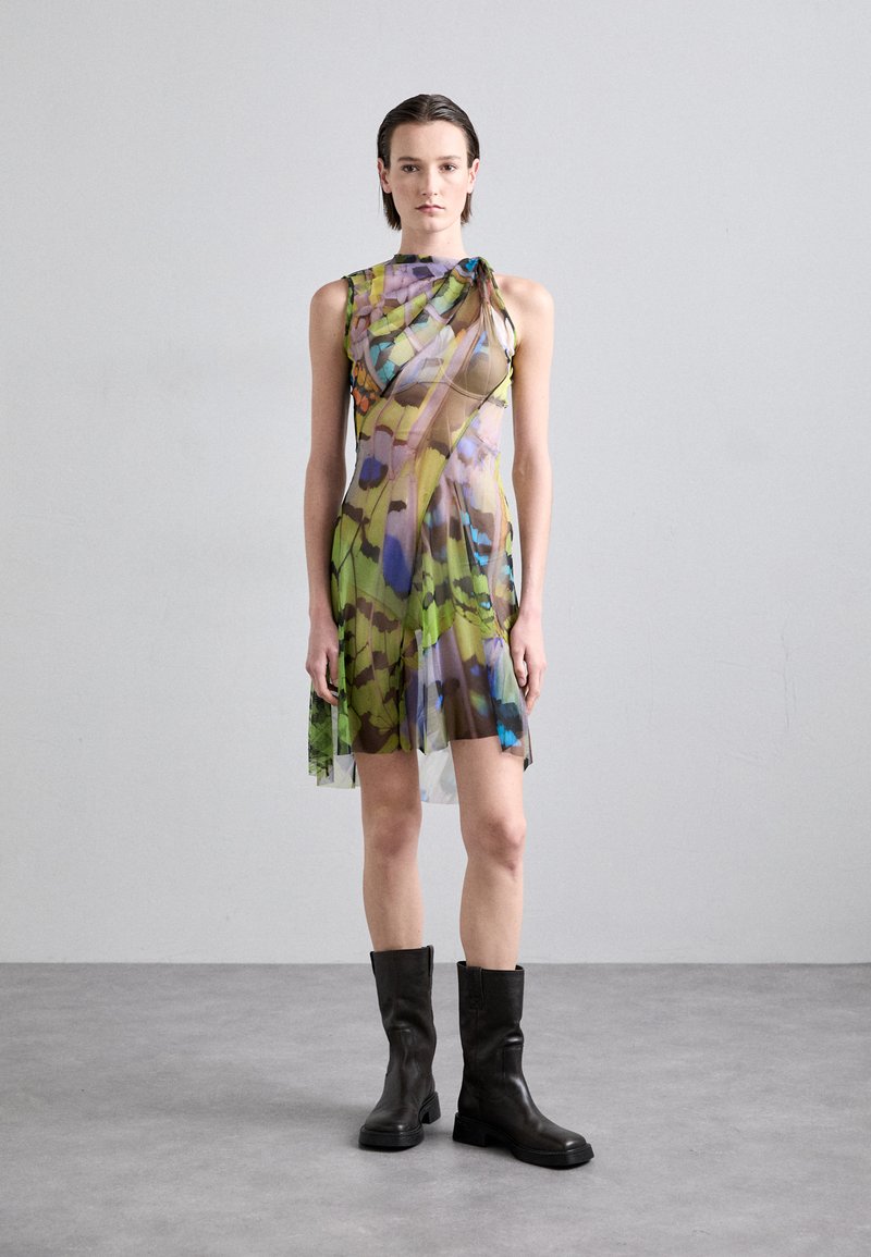 Robe multicolore avec une superposition transparente, présentant un motif floral dans des teintes de vert, bleu et brun. Associée à des bottes noires jusqu'aux genoux.