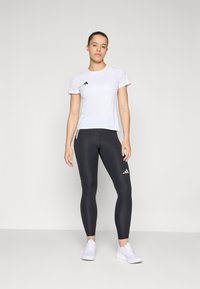Weißes Sport-T-Shirt mit schwarzen Akzenten, kombiniert mit schwarzen Leggings aus strukturiertem Stoff und seitlichen Streifen. Die Schuhe sind weiße Sneakers.