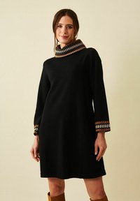 Nainen, jolla on päällään musta pitkähiha dressi, jossa on kuvioidut hihansuut ja korkeakauluksinen, yhdistettynä ruskeisiin polvipitkiin saappaisiin, seisoo yksinkertaisen taustan edessä.