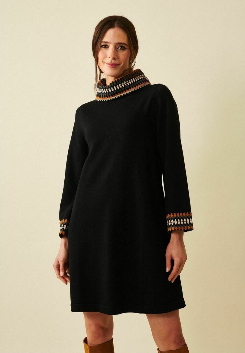 Nainen, jolla on päällään musta pitkähiha dressi, jossa on kuvioidut hihansuut ja korkeakauluksinen, yhdistettynä ruskeisiin polvipitkiin saappaisiin, seisoo yksinkertaisen taustan edessä.