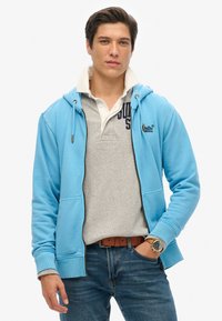 Sudadera de cremallera azul claro con cordones, polo gris debajo, vaqueros azules y cinturón marrón. Presenta un logo en el polo y un tejido texturizado.