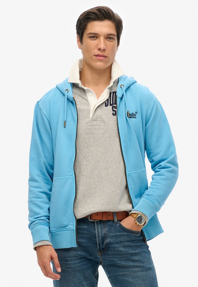 Sudadera de cremallera azul claro con cordones, polo gris debajo, vaqueros azules y cinturón marrón. Presenta un logo en el polo y un tejido texturizado.