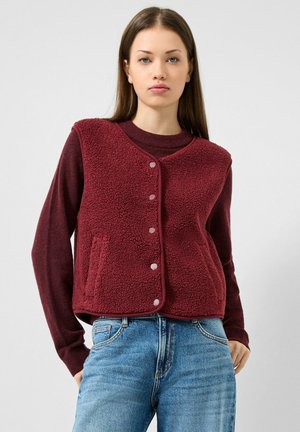 Giovane donna che indossa un gilet in pile bordeaux con bottoni sopra una maglia a maniche lunghe abbinata e jeans blu, in piedi su uno sfondo chiaro e uniforme.