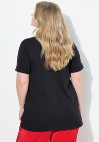 Czarna koszulka z krótkim rękawem o luźnym kroju, wykonana z miękkiego materiału, zestawiona z żywymi czerwonymi spodniami sztruksowymi. Widoczne długie, falowane włosy.