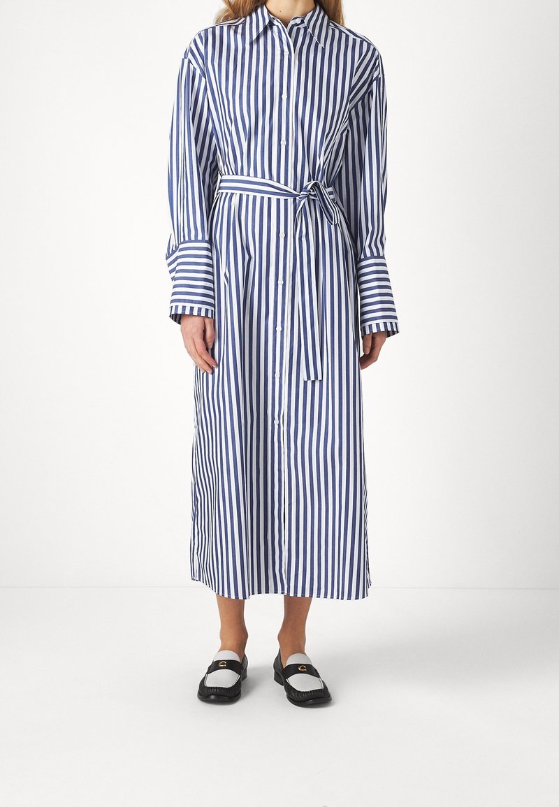 Femme portant une longue robe boutonnée à rayures verticales bleues et blanches avec une ceinture nouée à la taille et des mocassins noir et blanc.