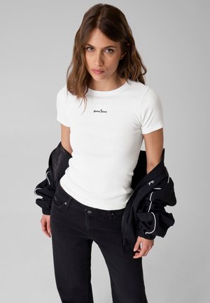 Mladá žena s hnědými vlasy, která má na sobě bílé tričko Guess Jeans a černou bundu přehozenou přes ramena na jednoduchém pozadí.