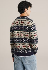 Suéter de punto con una base crema y patrones en azul marino, rojo y verde, incluyendo renos y copos de nieve; dobladillo y puños acanalados, cuello redondo.