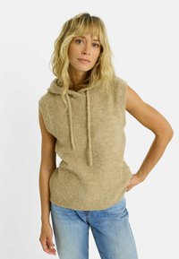 Pull à capuche sans manches beige en tissu doux et texturé. Dispose d'un cordon de serrage au cou et d'une coupe décontractée. Associé à un jean bleu clair.