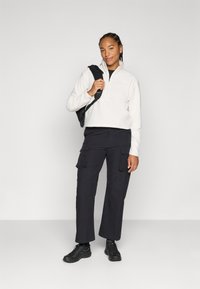 Pullover in pile color crema con colletto a mezza zip e logo; abbinato a pantaloni cargo neri con tasche; sneakers nere.