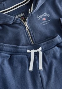 Sweat à capuche bleu Superdry avec zip et pantalon assorti doté d’un cordon blanc noué en nœud à la taille.
