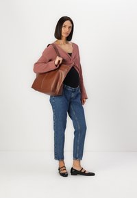 Braune Ledertasche mit glatter Textur, getragen von einer Person in einer weichen rosa Strickjacke und blauen Jeans. Schwarze Ballerinas runden das Outfit ab.
