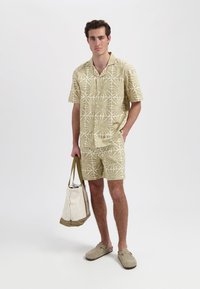 Beige gestreepte, kortemouwen, knoopsluiting shirt en korte broek, met een bloemdessin in een lichtere tint. Draagt een crèmekleurige draagtas met een bruine rand.