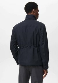 Hombre con chaqueta oscura de cuello alto y cintura ajustada, mostrado desde la parte de atrás contra un fondo claro y liso.