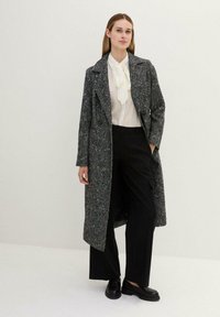 Cappotto lungo e strutturato in tessuto intrecciato bianco e nero, con colletto a bavero, design doppiopetto e tasche laterali, indossato sopra una blusa color crema e pantaloni neri.