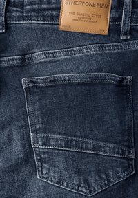 Mörkblå denimjeans med en bakficka, sömdetaljer och en tan läderetikett med varumärkestryck nära midjan.