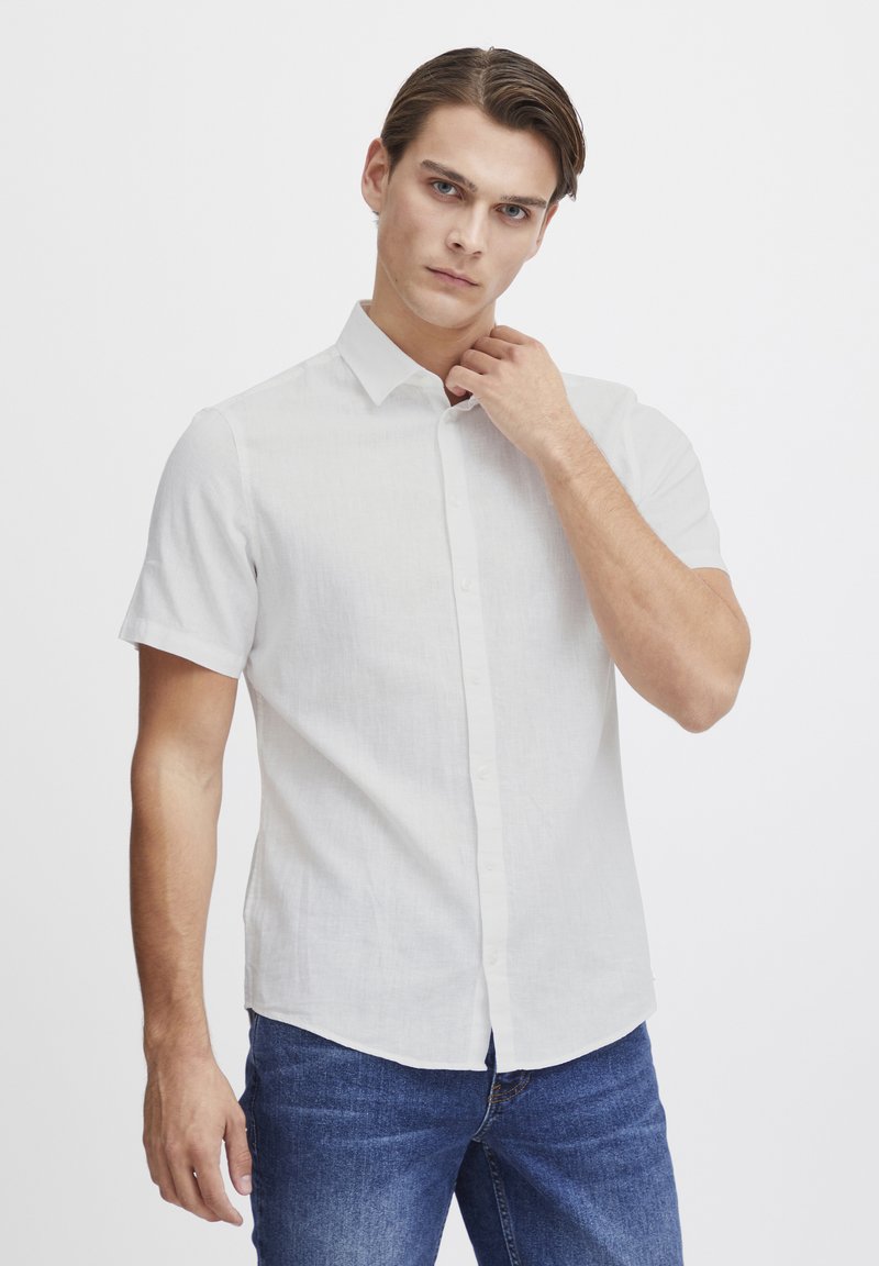Camisa blanca de manga corta con botones en tejido ligero. Textura suave, diseño con cuello y un ligero brillo. Llevada con jeans de mezclilla azules.