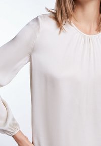 Blusa beige chiaro realizzata in tessuto liscio, con scollatura plissettata e maniche ampie con polsini elasticizzati. Design minimalista con linee semplici.