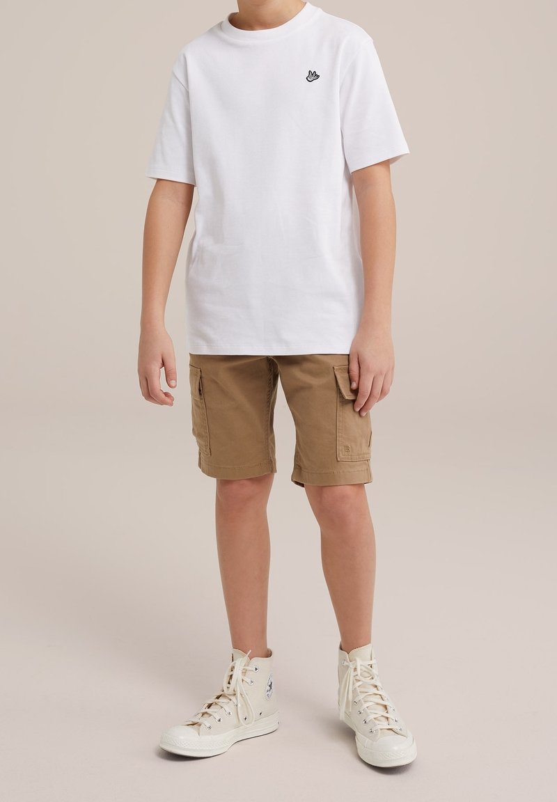 Enfant portant un t-shirt blanc avec un petit logo noir en forme de main, un short cargo marron et des baskets blanches montantes, debout devant un fond uni.