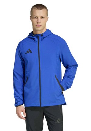 Un homme porte une veste Adidas bleue à capuche et à fermeture éclair sur le devant, avec un pantalon noir, sur fond blanc.