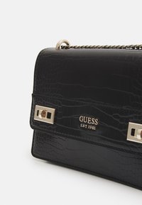 Černá kabelka s krokodýlím reliéfem, vybavená stříbrným řetízkem a dekorativními doplňky. Ozdobena logem „GUESS EST 1981“.