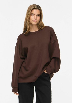 Kvinde med langt lys brun hår iført en løs mørkebrun sweater og sorte bukser, stående mod en ensfarvet hvid baggrund.