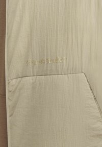 Veste beige clair avec une surface texturée, dotée d'une poche avant marquée à gauche et d'un logo "originals Studio™" dans une tonalité dorée subtile.